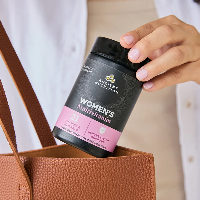 Hand holding a bottle of Ancient Nutrition Women's Multivitamin next to a brown bag.